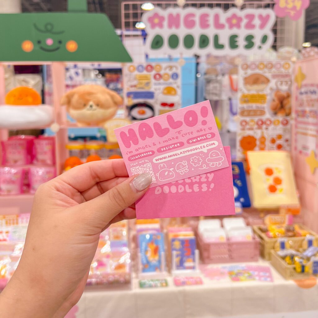 A hand is holding a unique business card in front of a colorful booth at a convention. The business card is pink with cute illustrations and reads "Hallo! I'm Angel & I make cute art." The background displays various products and a sign that says "Angelazy Doodles!" The booth is decorated with plush toys, stickers, and other merchandise, creating a vibrant and inviting atmosphere.