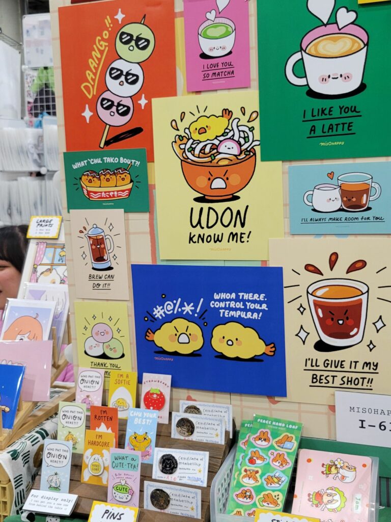 A colorful display at an Anime Expo Artist Alley table featuring various illustrated prints and merchandise. The prints include playful food-themed designs with puns such as "Udon know me!" with a bowl of noodles, "I like you a latte" with a cup of coffee, and "Control your tempura!" with tempura characters. The booth also showcases enamel pins, stickers, and certificates of membership. The setup is vibrant and eye-catching, with neatly arranged items and a variety of cute, whimsical artwork.