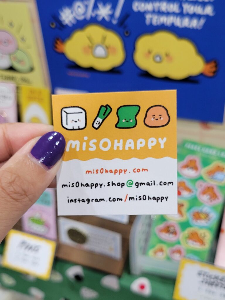  hand with purple-painted nails is holding a unique business card for "misOhappy" in front of a colorful booth background. The business card has a yellow and white design with cute illustrations of tofu, a green onion, seaweed, and a rice ball. It includes the website "misohappy.com," an email address "misohappy.shop@gmail.com," and the Instagram handle "instagram.com/mis0happy." The background shows various colorful products and signs displayed at a convention booth, creating a lively and inviting atmosphere.