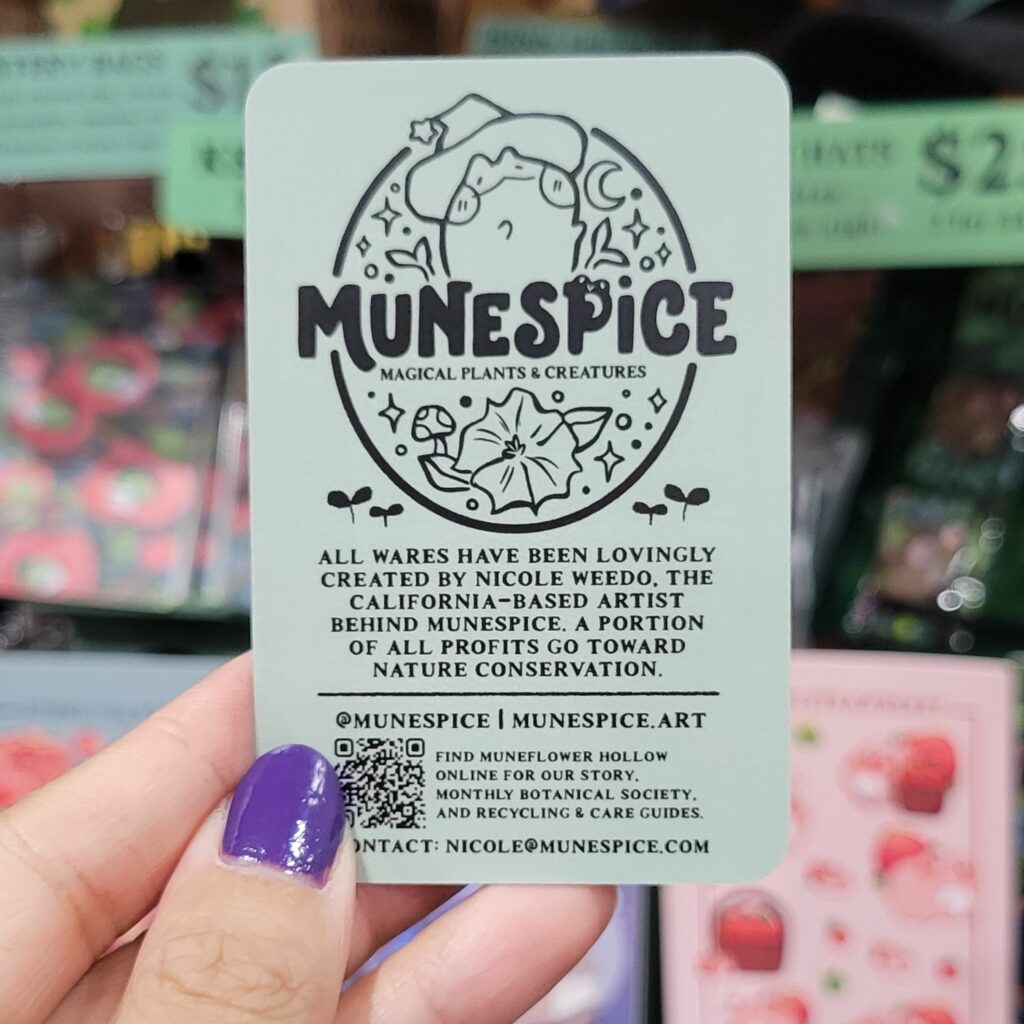 A hand with purple-painted nails is holding a unique business card for "MuneSpice," which features a cute illustration of a magical plant creature wearing a wizard hat. The card has a light green background and black text. It reads "MuneSpice - Magical Plants & Creatures" and includes a description stating that all wares are created by Nicole Weedo, with a portion of profits going toward nature conservation. The card also provides social media handles, a QR code, and contact information. The background shows various colorful products displayed at an Anime Expo Artist Alley table.