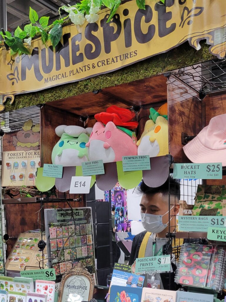 The Artist Alley table at Anime Expo featuring MuneSpice, a vendor specializing in "magical plants and creatures." The display includes plush toys shaped like tea frogs, rose frogs, and pumpkin frogs. Various items such as sticker sheets, postcard prints, bucket hats, keychain plushes, and mystery bags are neatly organized on shelves. The booth is decorated with green foliage and a sign that reads "MuneSpice - Magical Plants & Creatures." A person wearing a face mask stands behind the counter, ready to assist customers.