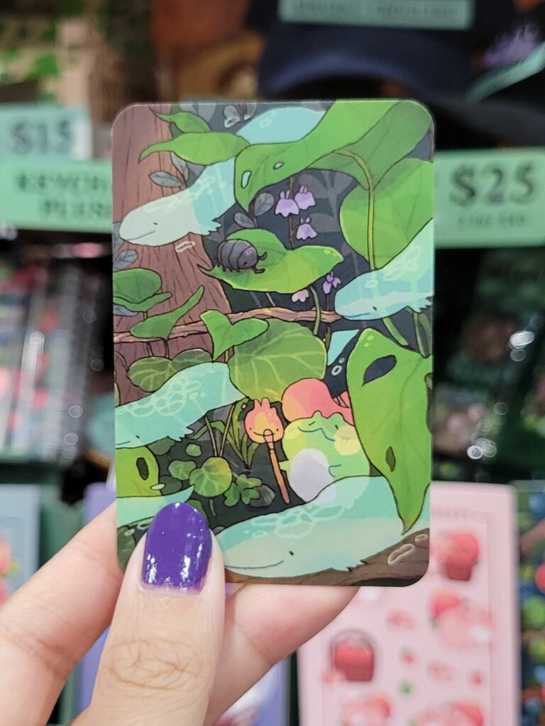  A hand with purple-painted nails is holding a business card featuring an illustration of a lush, whimsical forest scene. The artwork includes various plants, leaves, flowers, and a cute frog-like creature among the greenery. In the background, there are small, purple bell-shaped flowers and a roly-poly bug on a leaf. The scene is vibrant and detailed, capturing the enchanting atmosphere of a magical forest. The background of the image shows other items and signs displayed at the Munespice Artist Alley table at Anime Expo.