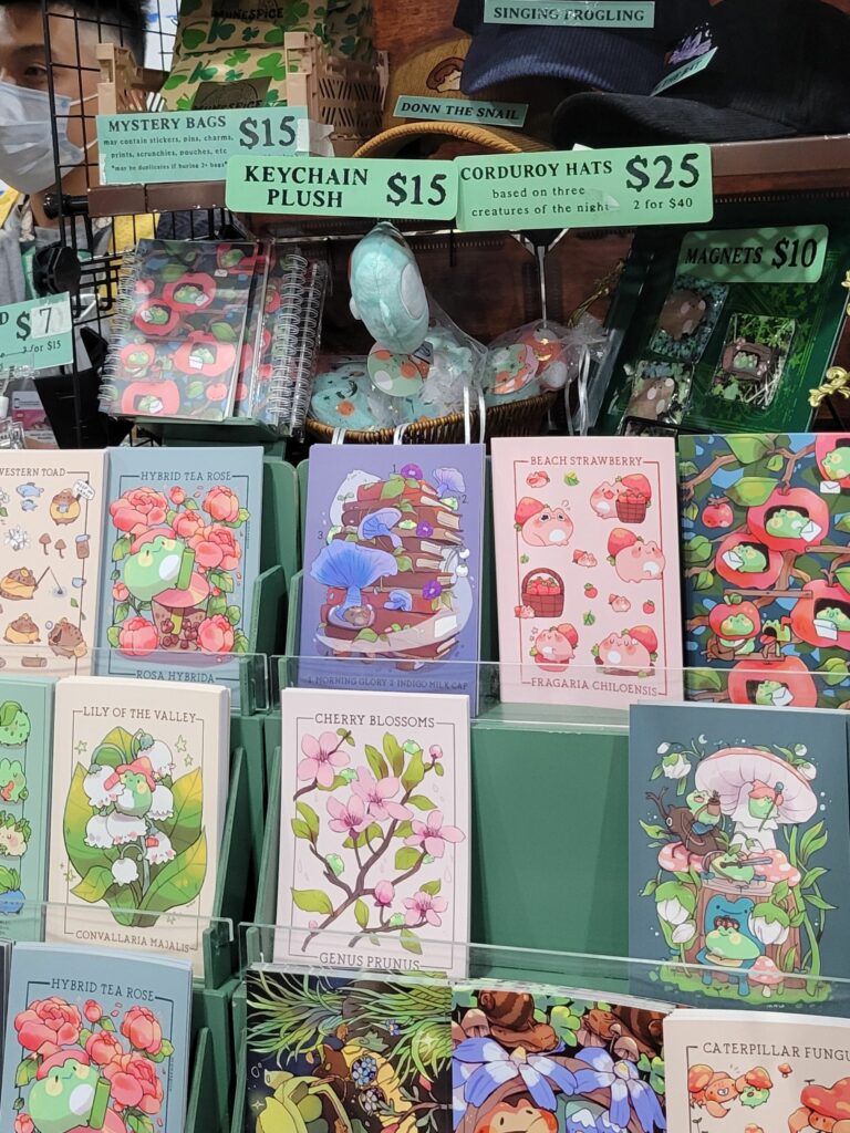 A display at Anime Expo featuring various illustrated prints, keychain plushes, and other merchandise from MuneSpice. The prints depict whimsical scenes of magical creatures, plants, and nature themes, including "Lily of the Valley," "Cherry Blossoms," and "Beach Strawberry." Signs indicate prices for items such as mystery bags ($15), keychain plushes ($15), corduroy hats ($25), and magnets ($10). The background shows a variety of colorful products and a vendor standing behind the display. The overall setup is vibrant and enchanting, highlighting the detailed and imaginative artwork.