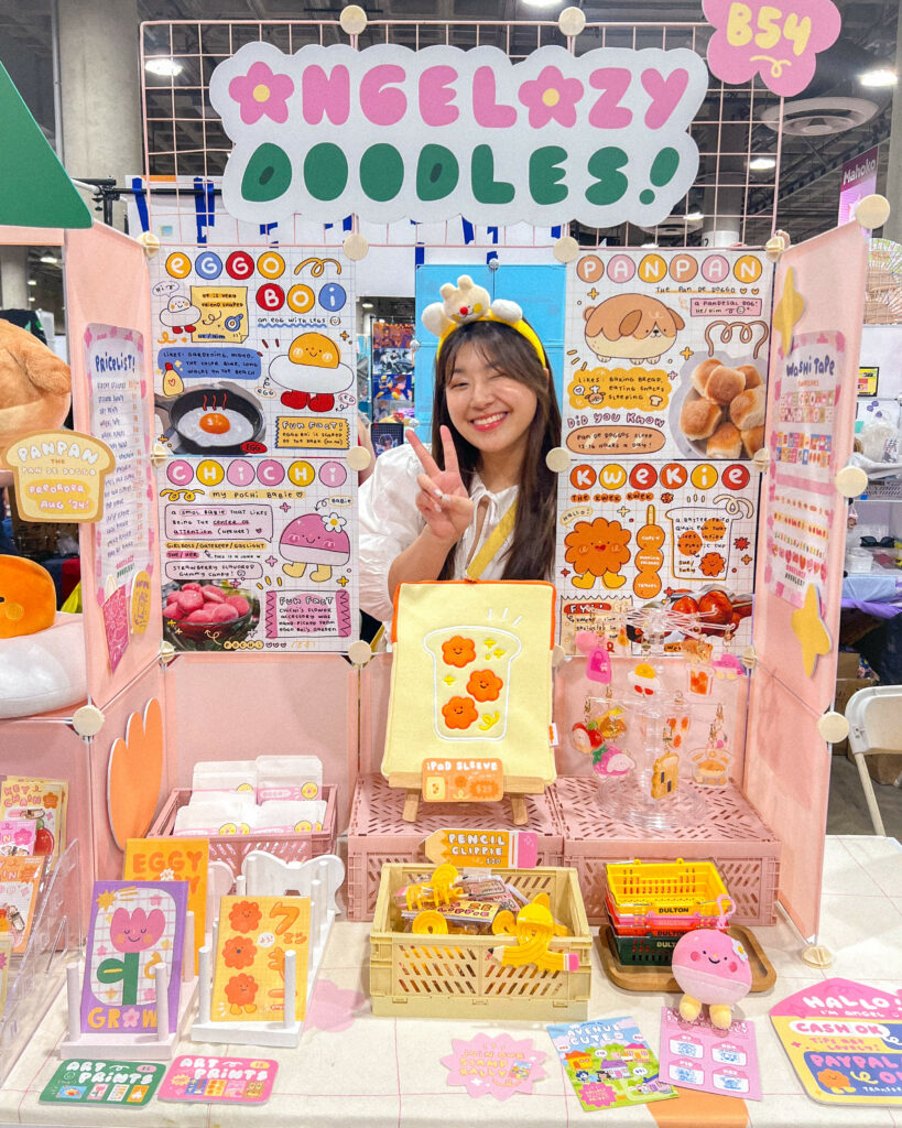 Angel, owner of Angelazy Doodles stands behind a colorful and well-organized booth at a convention, smiling and making a peace sign with her hand. The booth, labeled "Angelazy Doodles," displays a variety of cute and whimsical products including illustrations of eggs and other characters, plush toys, pencil clips, and an iPad sleeve with flower designs. The setup is vibrant with pink and yellow accents, and various items are neatly arranged on the table and shelves. The vendor is wearing a headband with cute decorations, matching the playful theme of the booth.