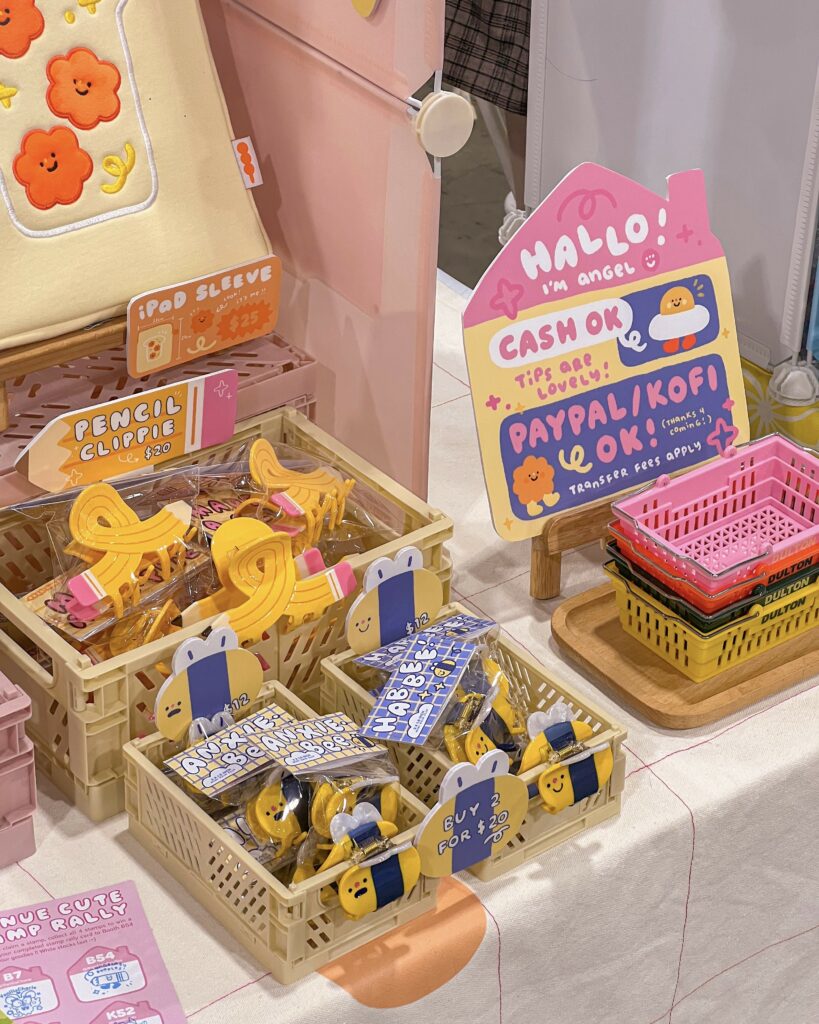 A display at an Anime Expo Artist Alley table featuring various cute and colorful products. The items include an iPad sleeve with flower designs, yellow pencil clips, and bee-themed hair clips. There is a sign that reads "Hallo! I'm Angel. Cash OK, tips are lovely! PayPal/Kofi OK!" with cute illustrations of characters. The products are neatly organized in small baskets, with additional colorful trays stacked on the side. The overall setup has a playful and inviting aesthetic, with pink and yellow accents.