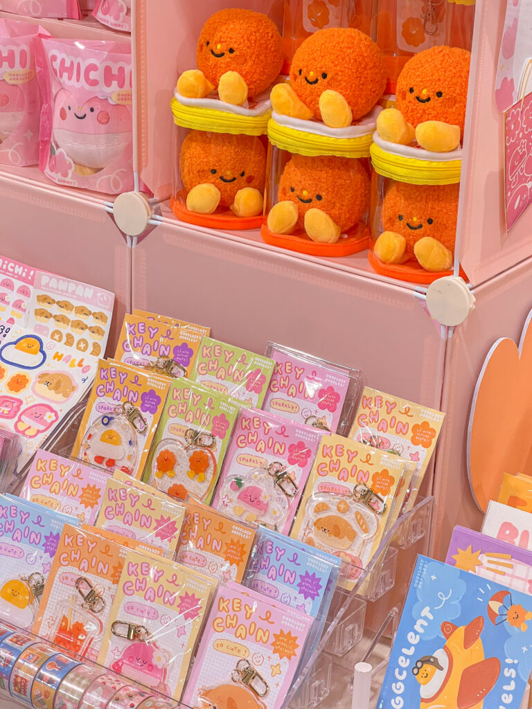 Part of the Angelazy Doodles Artist Alley display showcasing a variety of cute and colorful products. The upper section features plush toys of orange characters with smiling faces, seated in clear containers. Below them, several rows of keychains with various adorable designs are neatly arranged in clear holders. The keychains have colorful packaging in pink, yellow, green, and blue, featuring characters like eggs and other cute items. Additional products, such as stickers and decorative tapes, are visible at the bottom and sides of the display. The booth setup is bright and playful with a pink theme.