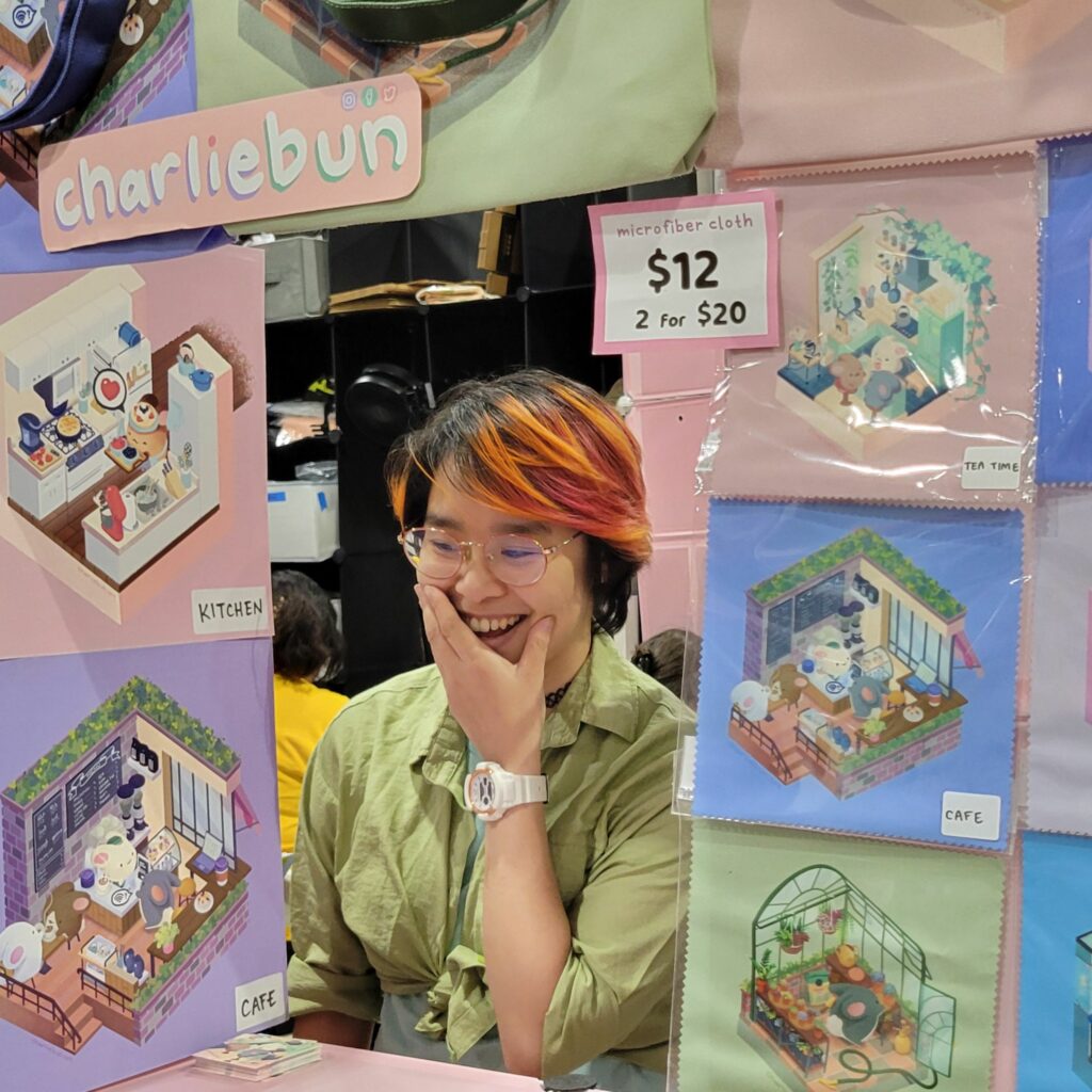 A person with glasses and colorful hair (orange and pink highlights) is smiling and covering their mouth with one hand while standing at a booth. The booth displays various colorful illustrations labeled "charliebun," including scenes of a kitchen, cafe, and other cute settings. There is a sign that reads "microfiber cloth $12, 2 for $20." The background shows a lively convention atmosphere with other attendees and booths visible.