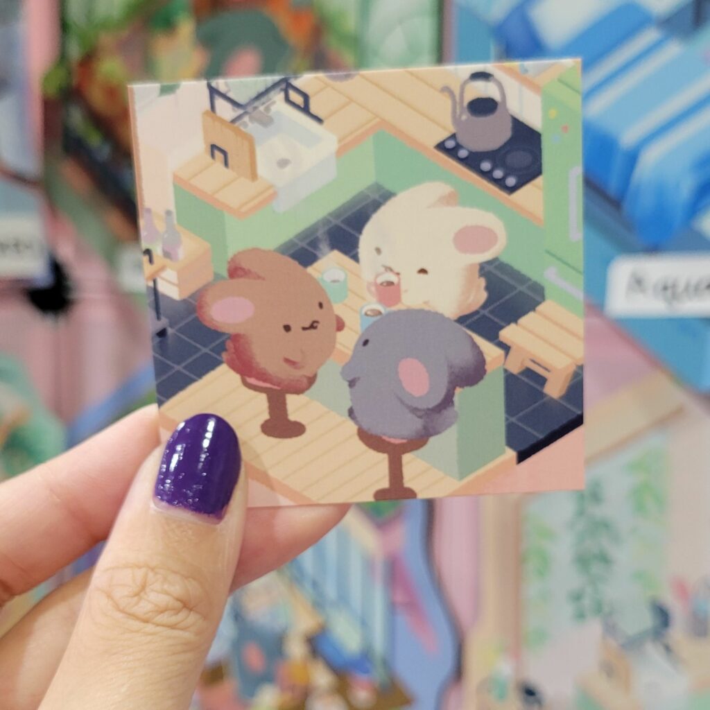 A hand with purple-painted nails is holding a unique business card featuring an adorable illustration of three cute bunnies sitting around a table in a cozy kitchen setting. The background shows various colorful illustrations or merchandise from a convention booth. The image on the business card is detailed and vibrant, highlighting the unique and charming design.