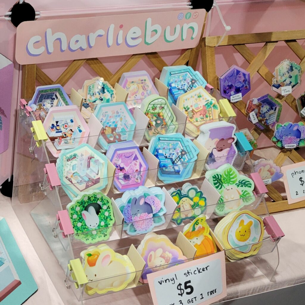  A display at an Artist Alley table at Anime Expo showcasing a variety of colorful vinyl stickers organized in a clear holder. The stickers feature cute and whimsical designs, including animals and cozy scenes. The booth is labeled "charliebun," and there is a sign that reads "vinyl sticker $5, buy 3 get 1 free." The setup includes multiple rows of hexagonal and other shaped stickers, with vibrant and detailed artwork. The overall display is inviting and neatly arranged.