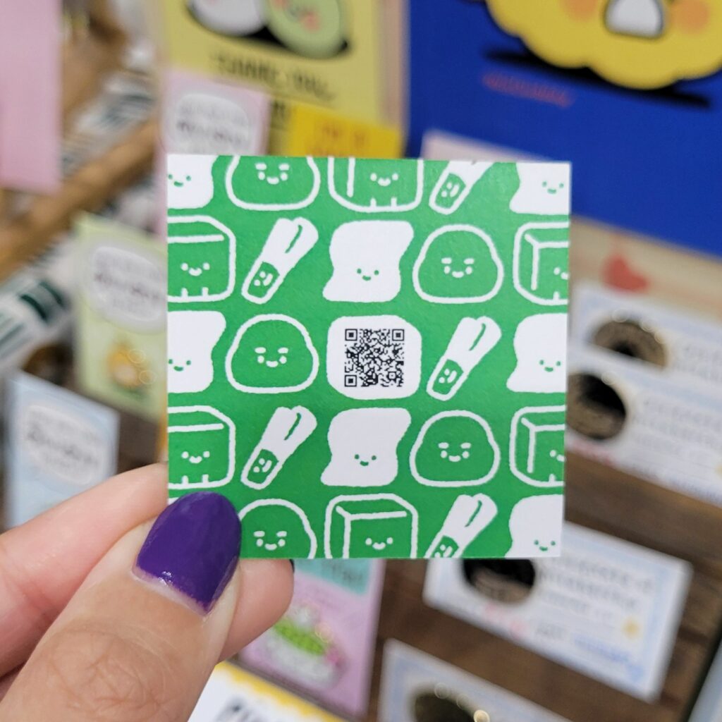 A hand with purple-painted nails is holding a unique business card with a green background and white illustrations of cute food characters, including smiling slices of bread and vegetables. In the center of the card, there is a QR code. The background of the image shows various colorful products displayed at a convention booth, adding to the lively atmosphere.