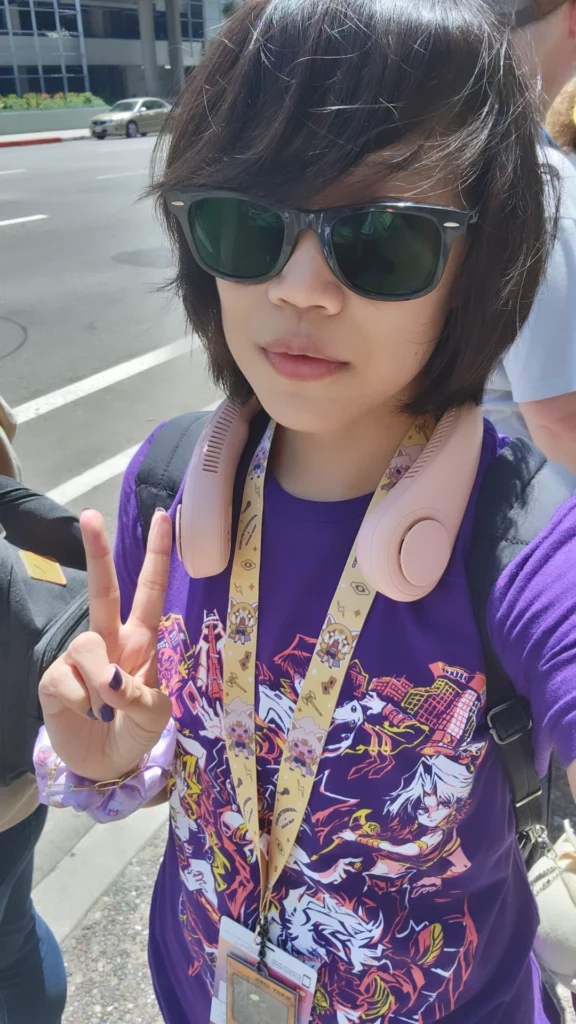 Designer Jess Macaraig wearing sunglasses and a purple graphic t-shirt is standing outside of the LA Convention Center. They have short, dark hair and are making a peace sign with their right hand. They are also wearing a lanyard with various badges and a pair of pink headphones around their neck. The background shows a street with a few cars and buildings.