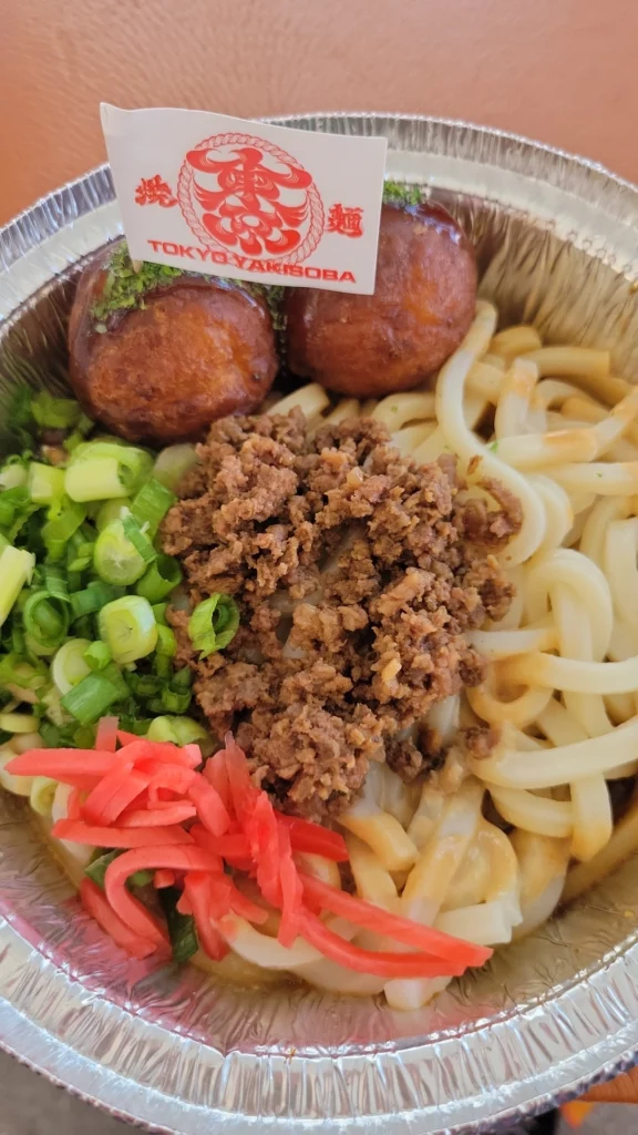 A close-up of a dish from Tokyo YakiSoba offered by the food trucks at Anime Expo. The dish includes thick noodles, ground meat, chopped green onions, pickled ginger, and two takoyaki balls. A small flag with the Tokyo YakiSoba logo is placed on top of the takoyaki balls. The food is served in a round aluminum container.