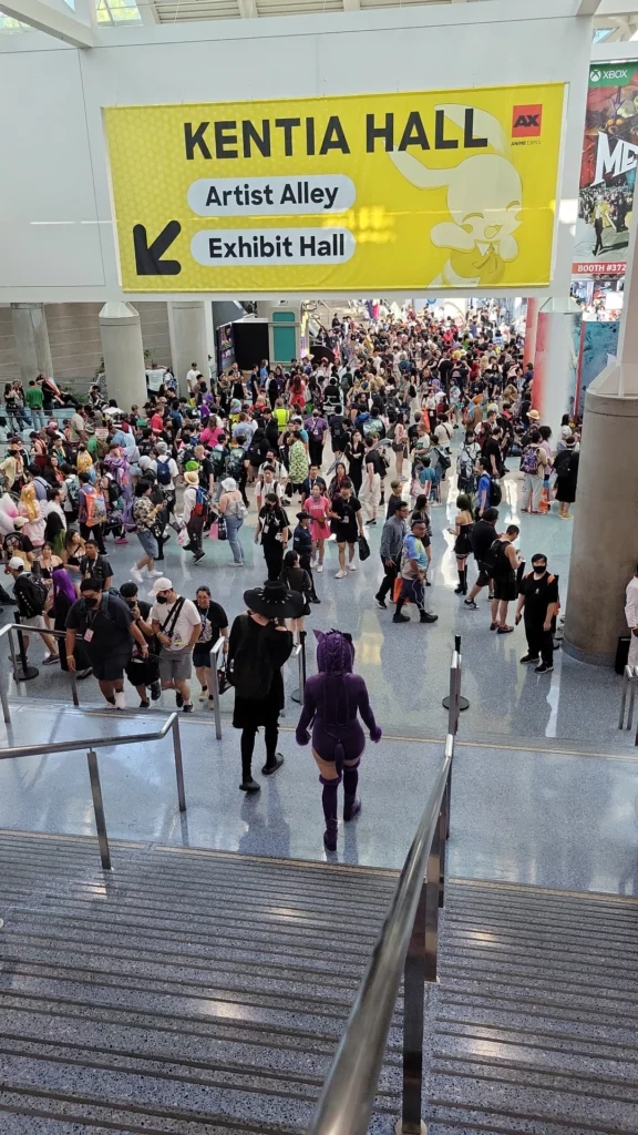 A large crowd of people at the LA Convention Center is seen from the top of a staircase. A yellow banner overhead reads "Kentia Hall" with directions to "Artist Alley" and "Exhibit Hall." Many attendees, some in cosplay, are walking and gathering in the spacious hall. A person in a purple costume is descending the stairs, and various booths and signs are visible in the background.