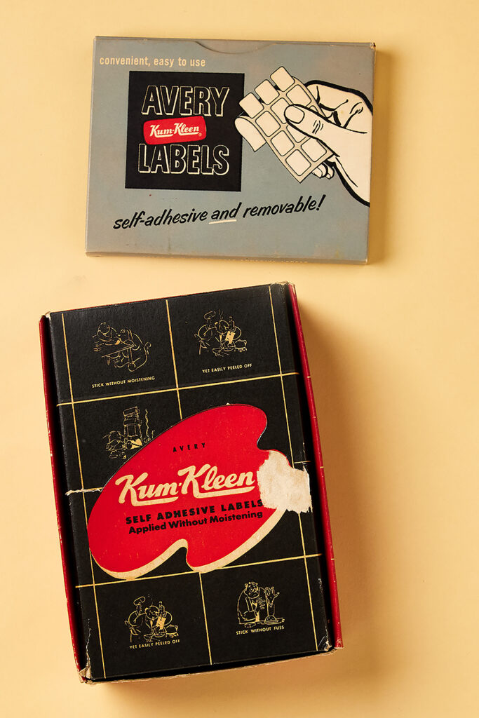 The image shows two vintage Avery label boxes, reflecting the history of stickers and adhesive labels. The top box has a retro design with a blue background, featuring an illustration of a hand peeling off small white rectangular labels. The text reads "Avery Kum-Kleen Labels" with the phrase "self-adhesive and removable!" below. The lower box has a black background with a red paint-shaped label and the text "Avery Kum-Kleen Self Adhesive Labels Applied Without Moistening." Surrounding the box are illustrations highlighting the ease of use, showing how the labels can be applied without moistening and easily peeled off, a key innovation in sticker history.




