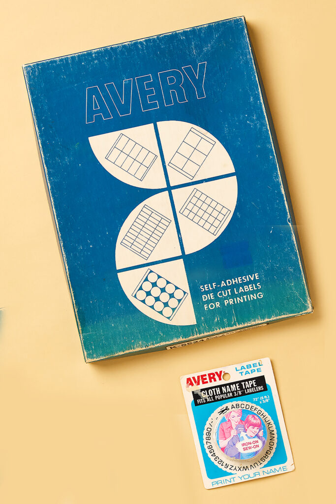 The image shows two vintage Avery packaging designs. The larger item is a blue box labeled "Avery Self-Adhesive Die Cut Labels for Printing," featuring simple white illustrations of various label shapes, such as rectangles and circles. The box has a worn texture, adding to its retro appeal. Below, there is a smaller package for "Avery Cloth Name Tape," which is designed for popular 3/8" labelers. The packaging features an illustration of a family with children, promoting its use for labeling clothes with iron-on or sew-on options. Both items reflect early innovations in labeling products.