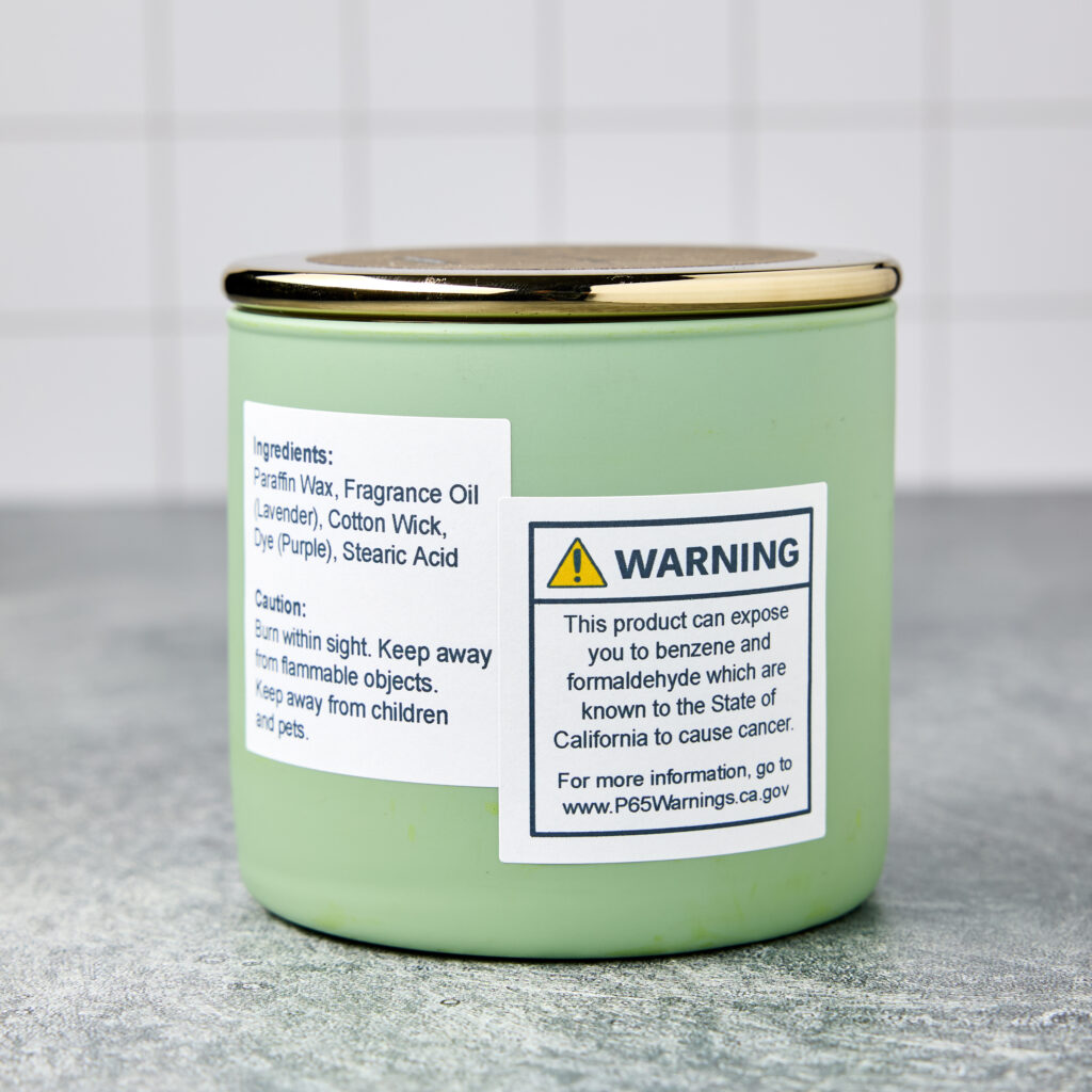 A green candle jar with a gold lid, featuring a Proposition 65 warning label designed with an Avery template and printed on a 1/2" square Avery TrueBlock label. The label blocks out any previous text and reads: "WARNING: This product can expose you to benzene and formaldehyde, which are known to the State of California to cause cancer. For more information, go to www.P65Warnings.ca.gov."