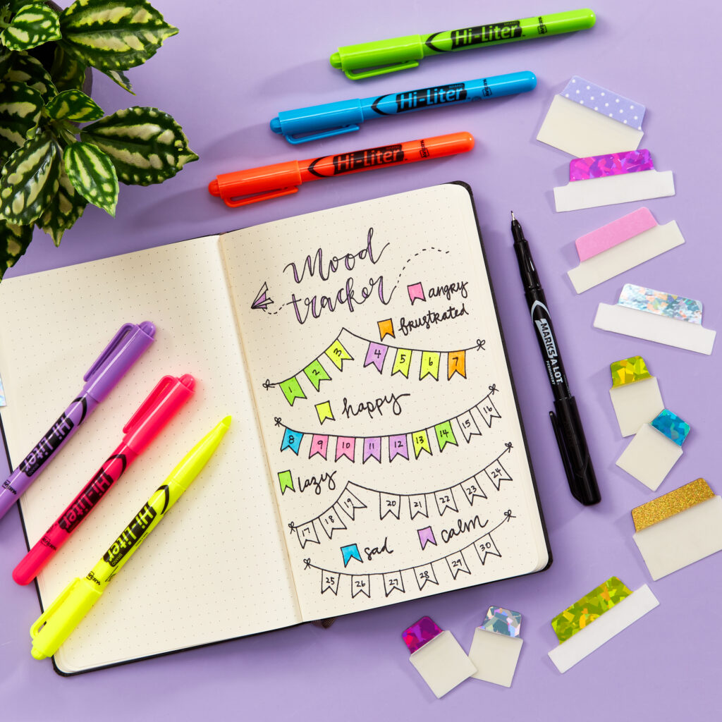 A bullet journal spread featuring a colorful mood tracker designed as hanging banners, each representing different emotions like angry, happy, lazy, sad, and calm. The layout is decorated with vibrant Hi-Liter markers and handwritten details for each day of the month. Surrounding the journal are Avery Ultra Tabs with glitter and patterns, a pen, and a potted plant. This creative setup serves as a fun and functional planner idea for tracking daily moods while adding artistic flair.