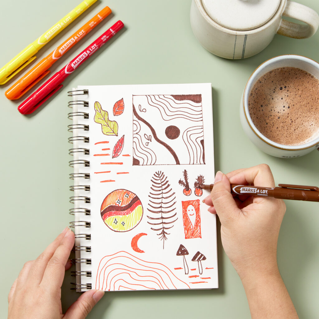 A person creates detailed and colorful sketches in a spiral-bound bullet journal using Avery Ultra Fine Tip Markers 9231 in yellow, orange, red, and brown. The artwork features abstract shapes, nature-inspired doodles, and whimsical designs. A warm cup of cocoa and a ceramic teapot sit nearby, adding a cozy touch to the journaling setup. The scene highlights creativity and mindfulness through journaling.