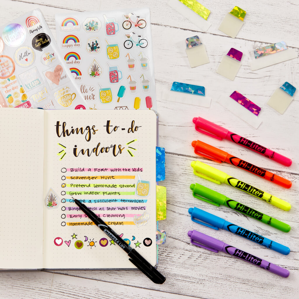 A flat lay featuring a planner open to a "Things to Do Indoors" list written in bold black text, decorated with colorful Avery 23585 highlighters, stickers, and vibrant washi tape tabs. The planner idea showcases creative indoor activities, such as building a fort, growing indoor plants, and making a succulent terrarium. Surrounding the planner are six Avery Hi-Liter markers in neon shades, sticker sheets with playful designs, and decorative tabs, offering inspiration for organizing and personalizing planners.