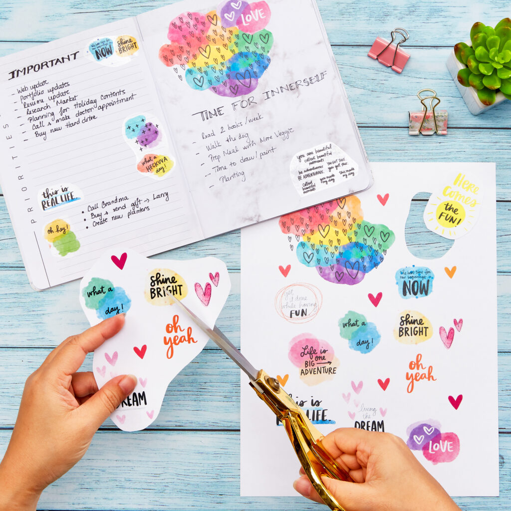 A person uses gold scissors to cut custom stickers from a sheet made with Avery 53202 sticker paper, featuring vibrant watercolor designs and motivational phrases like "Shine Bright" and "Oh Yeah." The planner in the background includes organized sections labeled "Important" and "Time for Inner Self," offering inspiration for a creative planner idea. The workspace is styled with colorful stationery, paper clips, and a small succulent, creating an inviting environment for planning and goal setting.