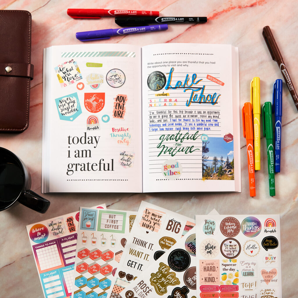 A gratitude journal spread decorated with Avery planner stickers featuring motivational quotes, colorful designs, and travel-themed elements. The layout includes prompts like "Today I am grateful" and reflections on a visit to Lake Tahoe, accented with handwritten notes and vibrant stickers. Surrounding the journal are sticker sheets, markers, and a coffee cup, offering a creative planner idea for expressing gratitude and capturing meaningful moments.