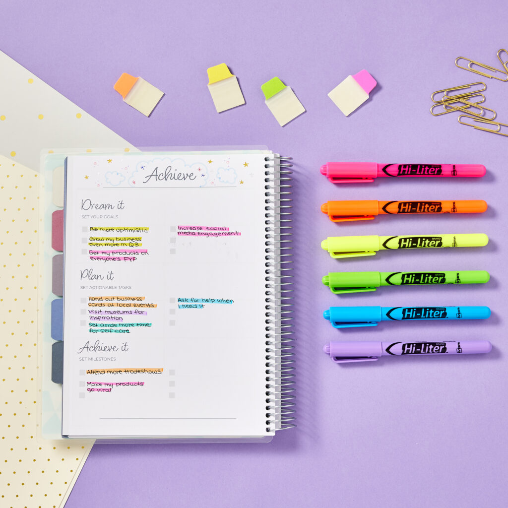 A spiral-bound notebook showcasing a goal-setting layout titled "Achieve," organized into sections for dreaming, planning, and achieving goals. Avery 23585 highlighters in vibrant colors are used to emphasize tasks and milestones, while Avery 74762 Ultra Tabs divide sections for easy navigation. Surrounding the notebook are additional Ultra Tabs, highlighters, paperclips, and a dotted folder, creating a colorful and functional setup for organizing goals and tasks.