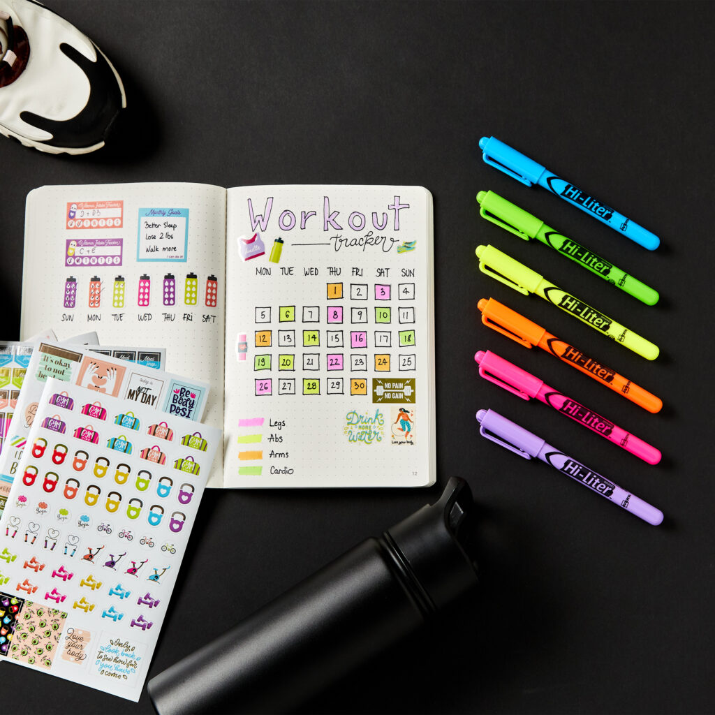 A workout tracker spread decorated with Avery 6787 fitness stickers and highlighted with Avery 23585 highlighters. The layout includes a calendar for tracking daily workouts, color-coded sections for different muscle groups, and motivational stickers. Surrounding the journal are fitness-themed sticker sheets, a reusable water bottle, and a pair of athletic shoes, showcasing a creative planner idea for staying organized and motivated with fitness goals.