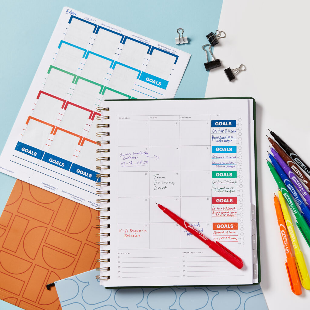 A monthly calendar spread featuring a free Avery template for goal stickers, organized with color-coded sections for tracking objectives. The layout includes handwritten notes for key events, like sales leadership and team building, alongside colorful goal labels for added focus. Surrounding the calendar are vibrant markers, binder clips, and extra sticker templates, showcasing a practical planner idea for organizing goals and deadlines efficiently.