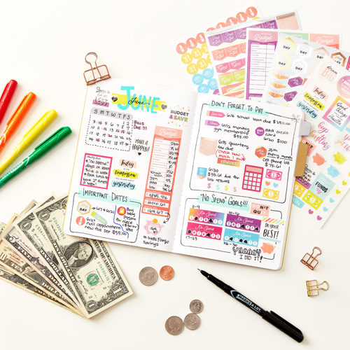 A bullet journal spread decorated with Avery 6788 budget stickers to track financial goals and monthly expenses. The pages include sections for a calendar, bill reminders, savings goals, and a "No Spend Goals" tracker. Vibrant stickers, colorful markers, and handwritten notes enhance the layout's functionality and visual appeal. Surrounding the journal are cash, coins, pens, and additional sticker sheets, providing a creative and organized approach to managing personal finances.