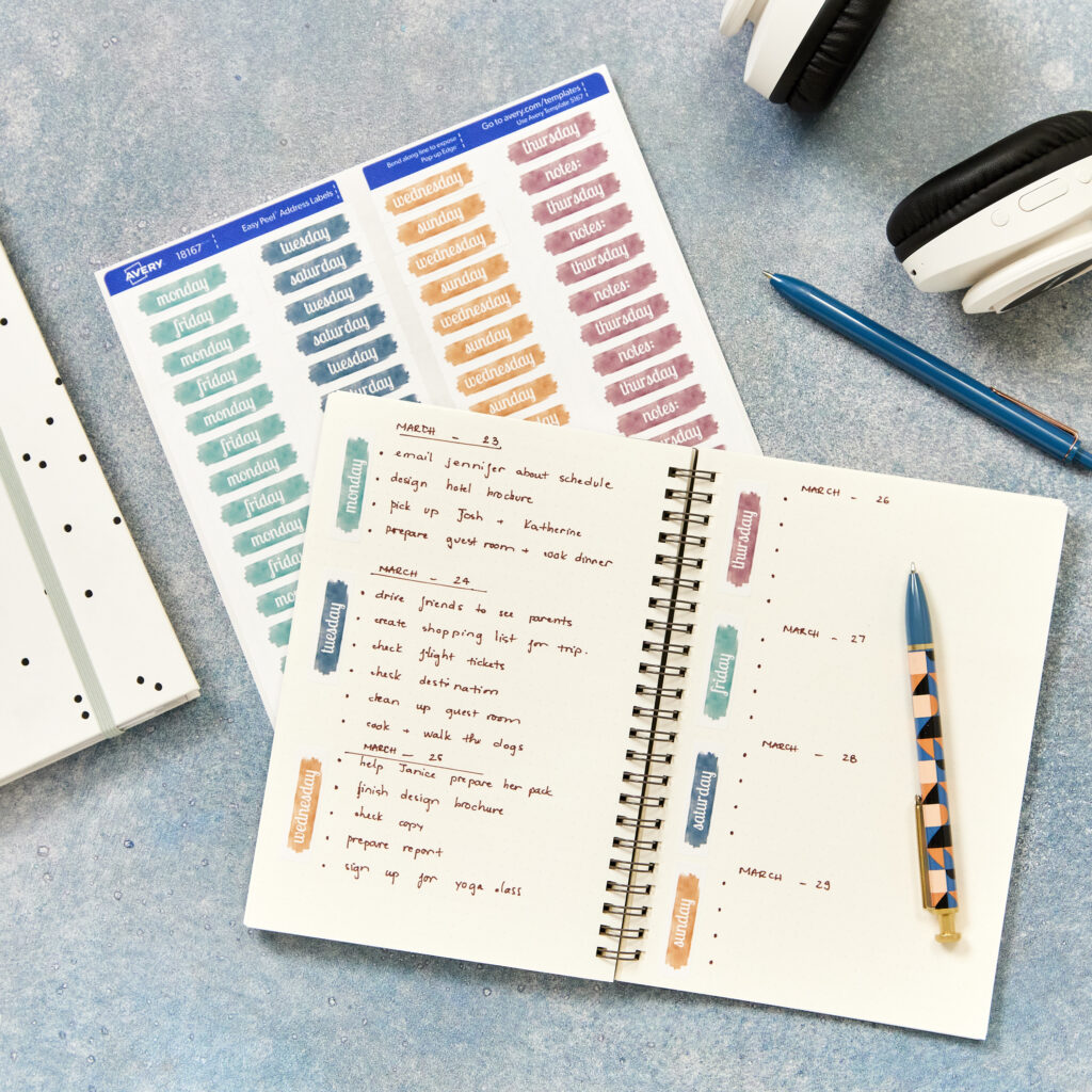 A neatly organized notebook with handwritten daily tasks enhanced by free Avery sticker designs featuring watercolor-style day-of-the-week labels. Surrounding the notebook are additional sticker sheets, a decorative pen, headphones, and a dotted binder.