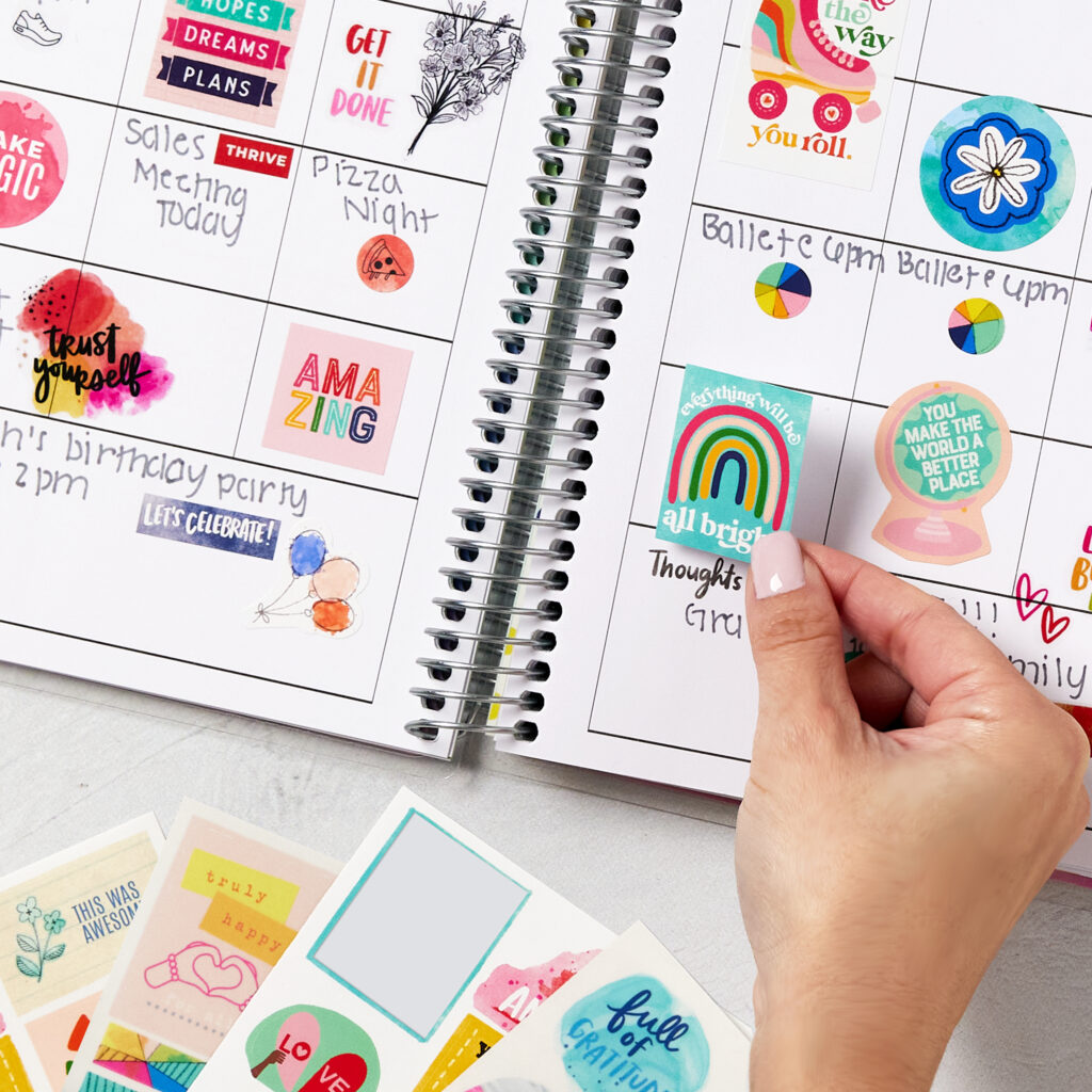 Calendar pages decorated with vibrant stickers from the Avery x Amy Tangerine collection, featuring motivational phrases, colorful designs, and playful icons like rainbows, balloons, and flowers. A hand is adding a sticker with the phrase "Everything will be all bright," enhancing a creative and personalized layout for tracking events and tasks. Additional sticker sheets are placed nearby, showcasing a variety of designs for customization and inspiration.